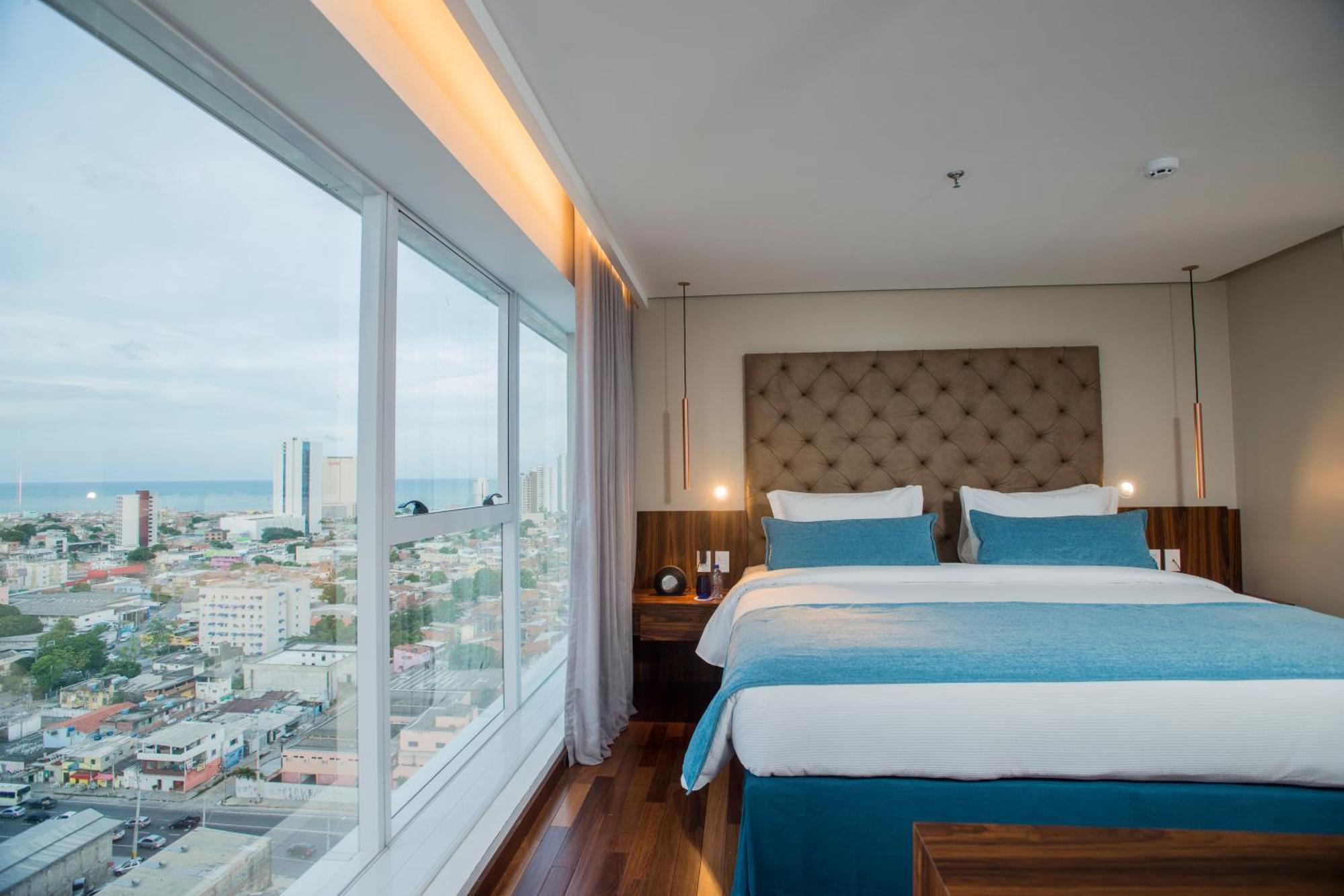Hotel Luzeiros Recife Exteriör bild The photo shows a modern hotel room featuring a large window with a city view. Inside, there are two neatly made beds with light blue and white bedding, flanked by wooden nightstands. The room has a contemporary design, with a padded headboard and so
